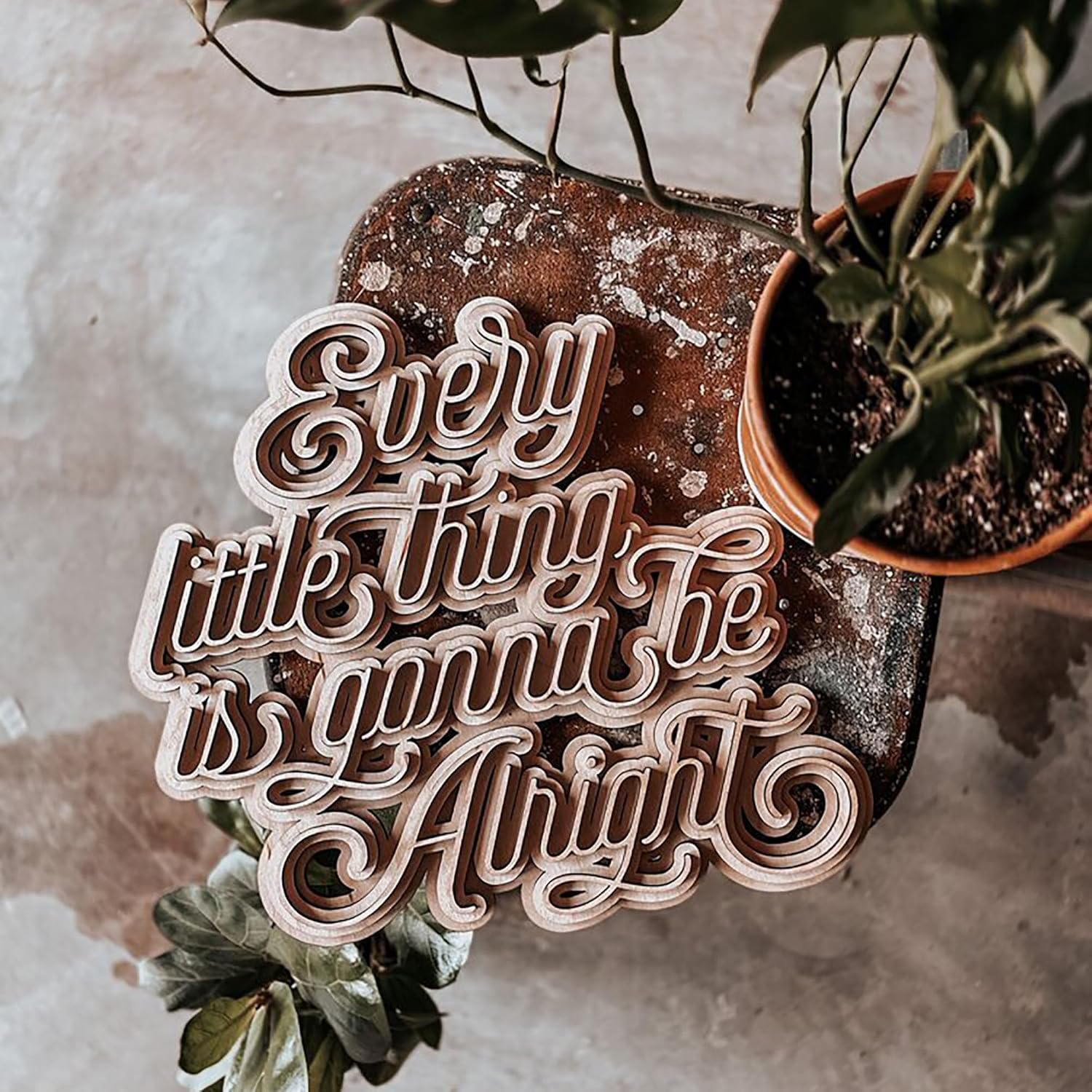 Wooden sign with 'Every little thing is gonna be alright' text on a textured surface with plants.