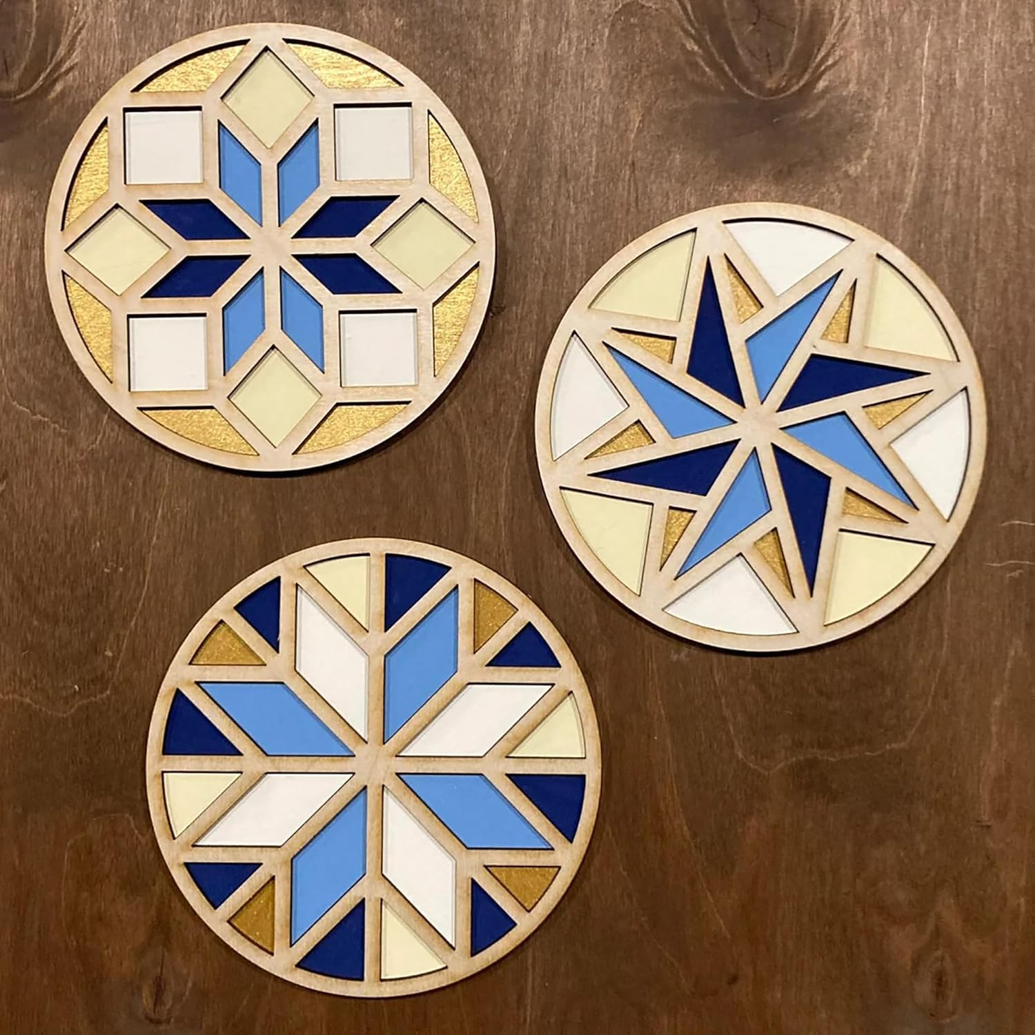 Three geometric coasters with blue, white, and gold colors on a wooden surface.