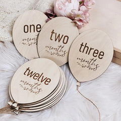 Wooden milestone markers for baby's first year with text 'one month', 'two months', 'three months', and 'twelve months' on a soft surface with flowers.