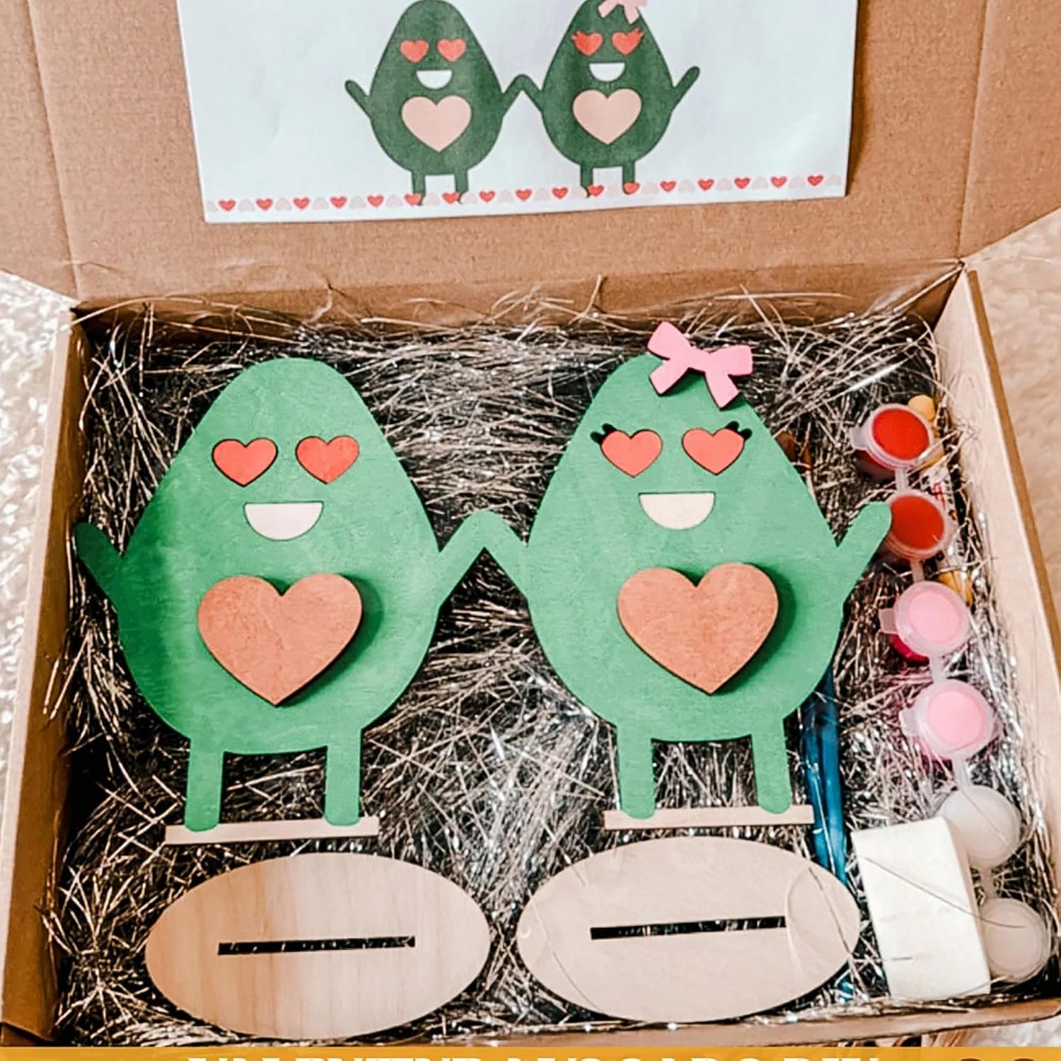 Two green wooden characters with hearts in a box with craft supplies