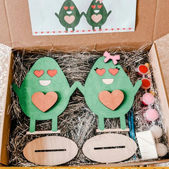 Two green wooden characters with hearts in a box with craft supplies