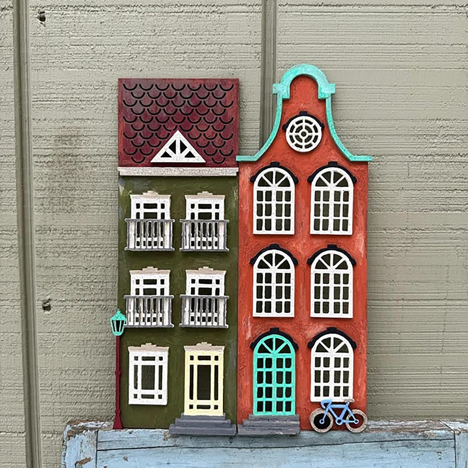 Decorative wall art of a house with windows and a bicycle on a textured wall.