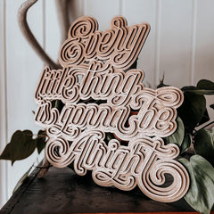 Wooden decorative sign with 'Every little thing is gonna be alright' text on a surface with plants.