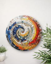 Colorful spiral mosaic wall art on a white wall with plants.