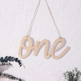 Wooden 'one' decoration hanging on a white textured wall with greenery at the bottom.