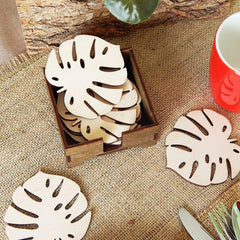 Wooden leaf coasters in a box on a textured surface with a red mug and green plant.