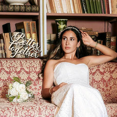 Woman in a white dress sitting on a patterned couch with a 'Better Together' sign and books in the background.