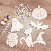 Halloween-themed wooden cutouts including a ghost, spider web, pumpkin, bat, cat, owl, and witch hat on a wooden surface with paint and brushes.