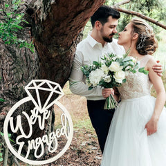 Engaged couple in a forest with 'We Are Engaged' sign.