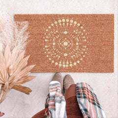 Doormat with gold mandala design on a light surface, next to dried plants and a person wearing colorful socks.