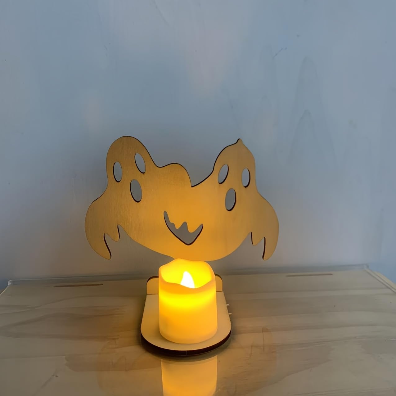 Wooden ghost-shaped candle holder with a lit candle on a light wooden surface.