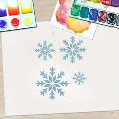 Blue snowflakes drawn on a white sheet of paper with watercolor paint palettes in the background.
