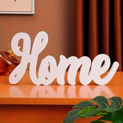 Decorative word 'Home' on a wooden shelf with a plant and framed picture in the background.