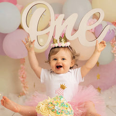Child celebrating a first birthday with balloons and a cake, wearing a crown and holding up the word 'one'.