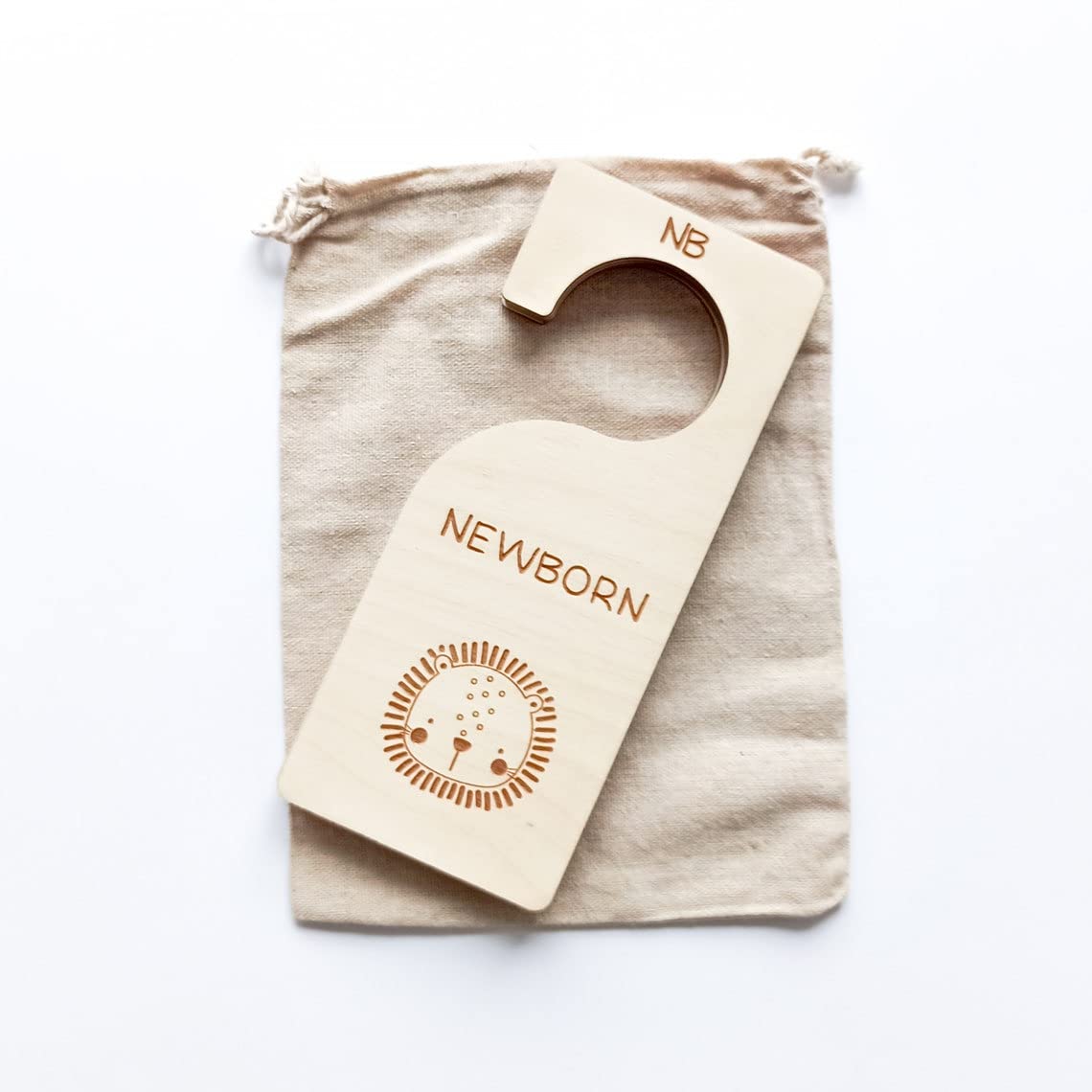 Wooden baby milestone card with 'NEWBORN' and 'NB' text on a beige drawstring bag.