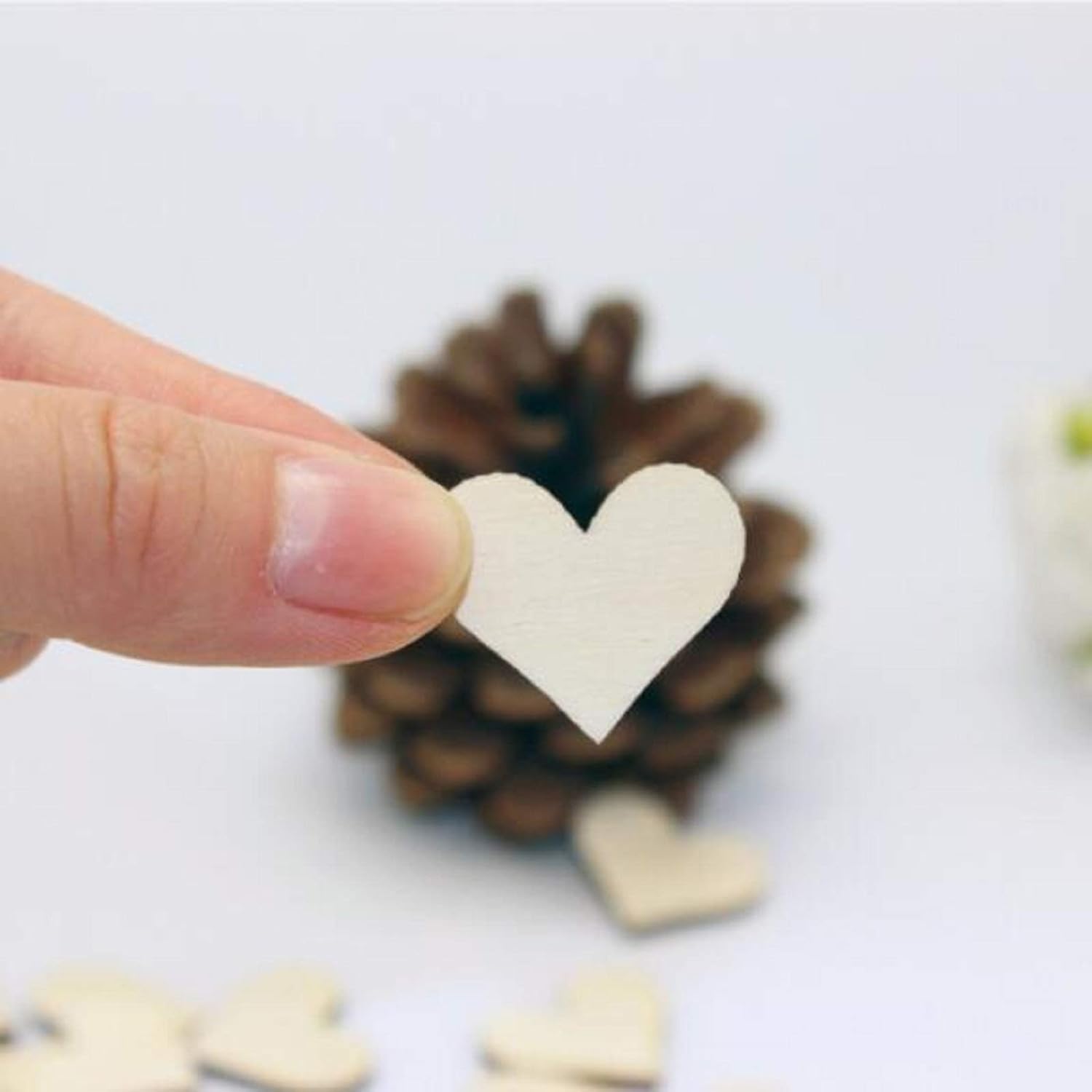 Hand holding a white heart-shaped object with blurred pinecones and more hearts in the background.
