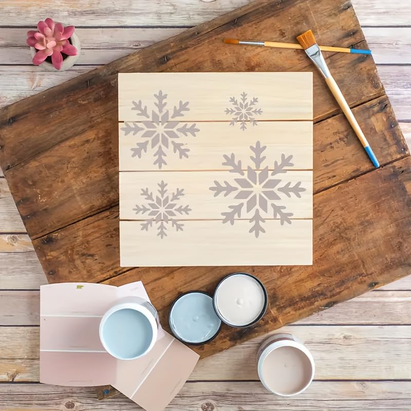 Wooden board with snowflake patterns, paintbrushes, and paint containers on a rustic wooden surface.
