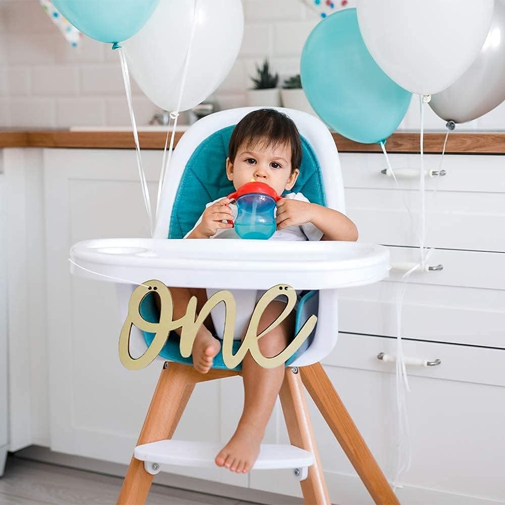 Child in a high chair with balloons and a 'one' sign, indicating a first birthday celebration.