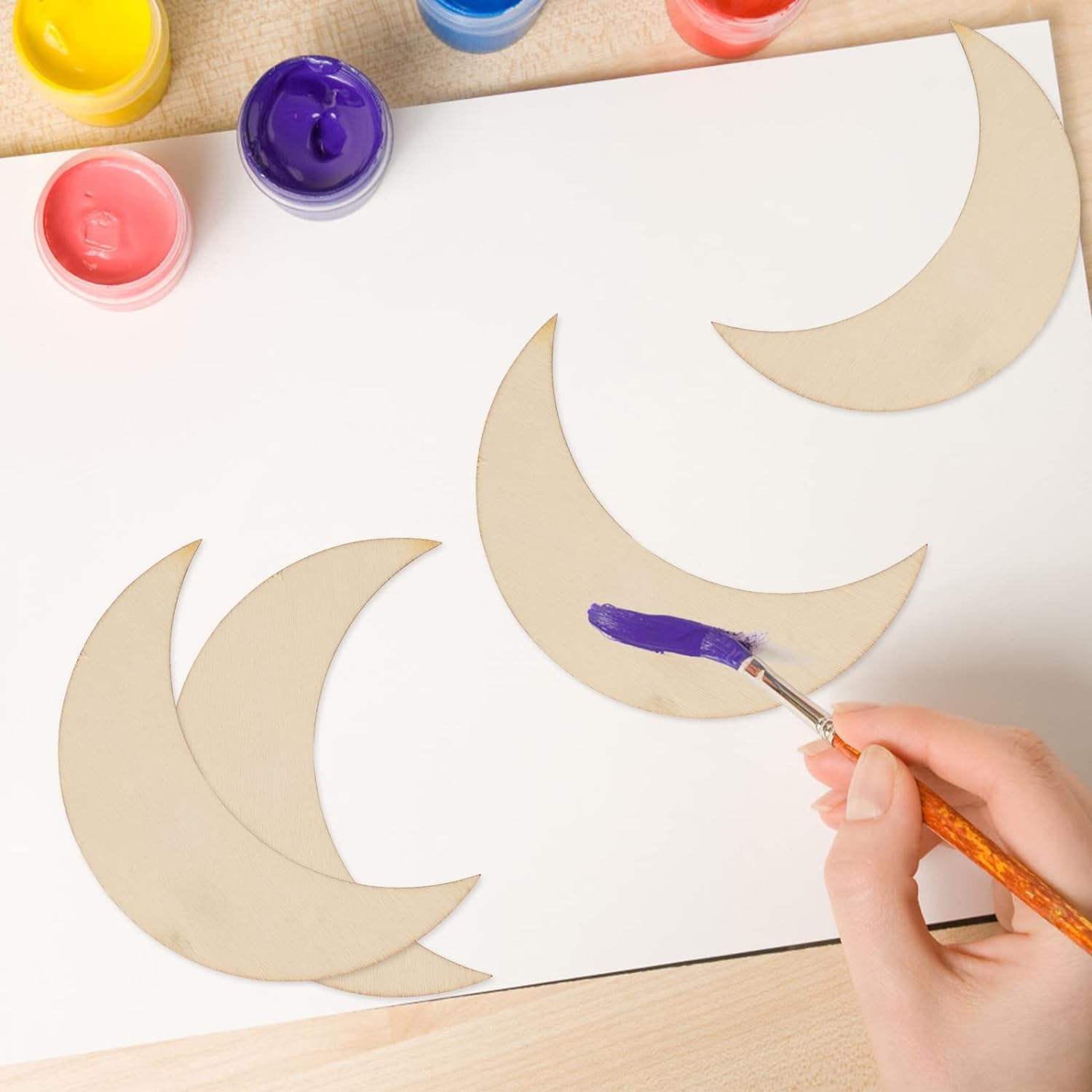 Person painting crescent moon shapes with a brush on a white sheet of paper, surrounded by colorful paint containers.