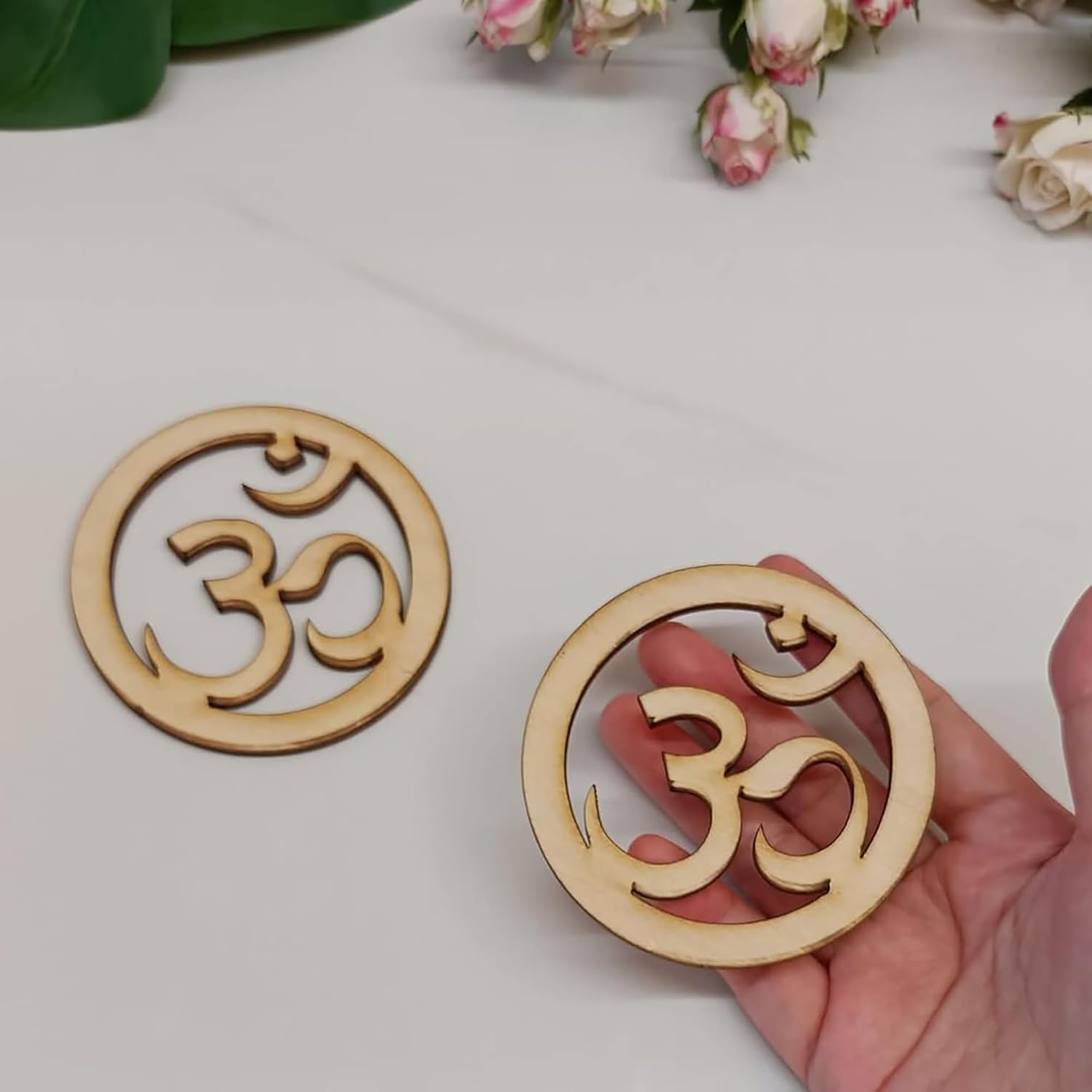 Wooden coasters with Om symbol held by a hand on a light surface with flowers in the background