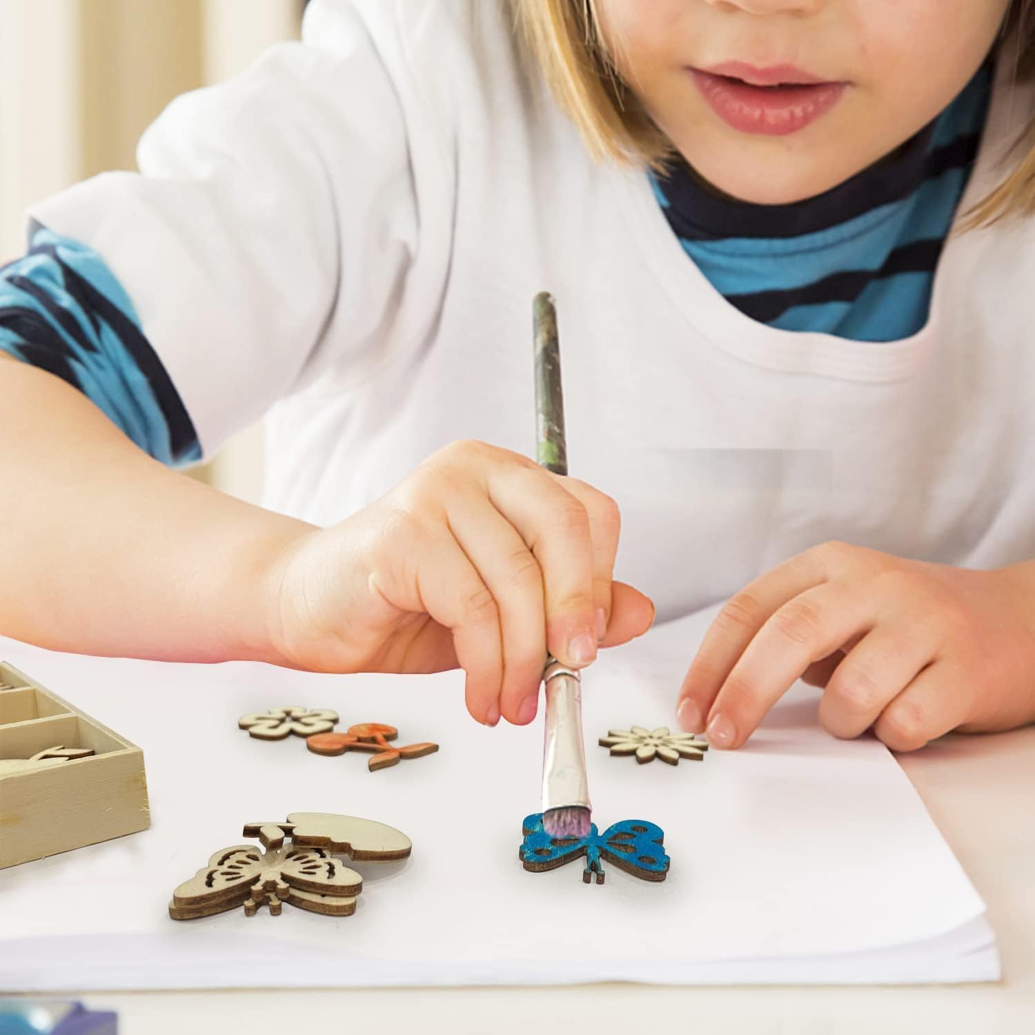 Child painting wooden butterfly figurines with a paintbrush on a white surface.