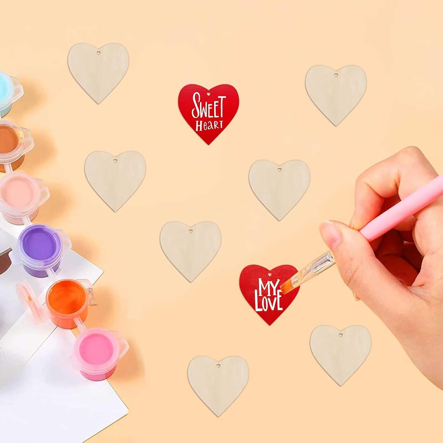 Person painting a red heart-shaped tag with 'My Love' text on a beige surface with paint containers and heart shapes.