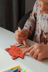 Child coloring a wooden craft with markers on a white table.