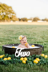 Baby in a bathtub with lemons and a 'Two' sign in a field