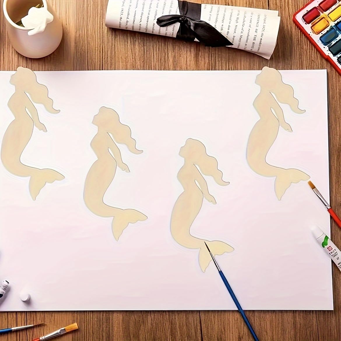 Mermaid silhouette cutouts on a piece of paper with art supplies on a wooden surface.