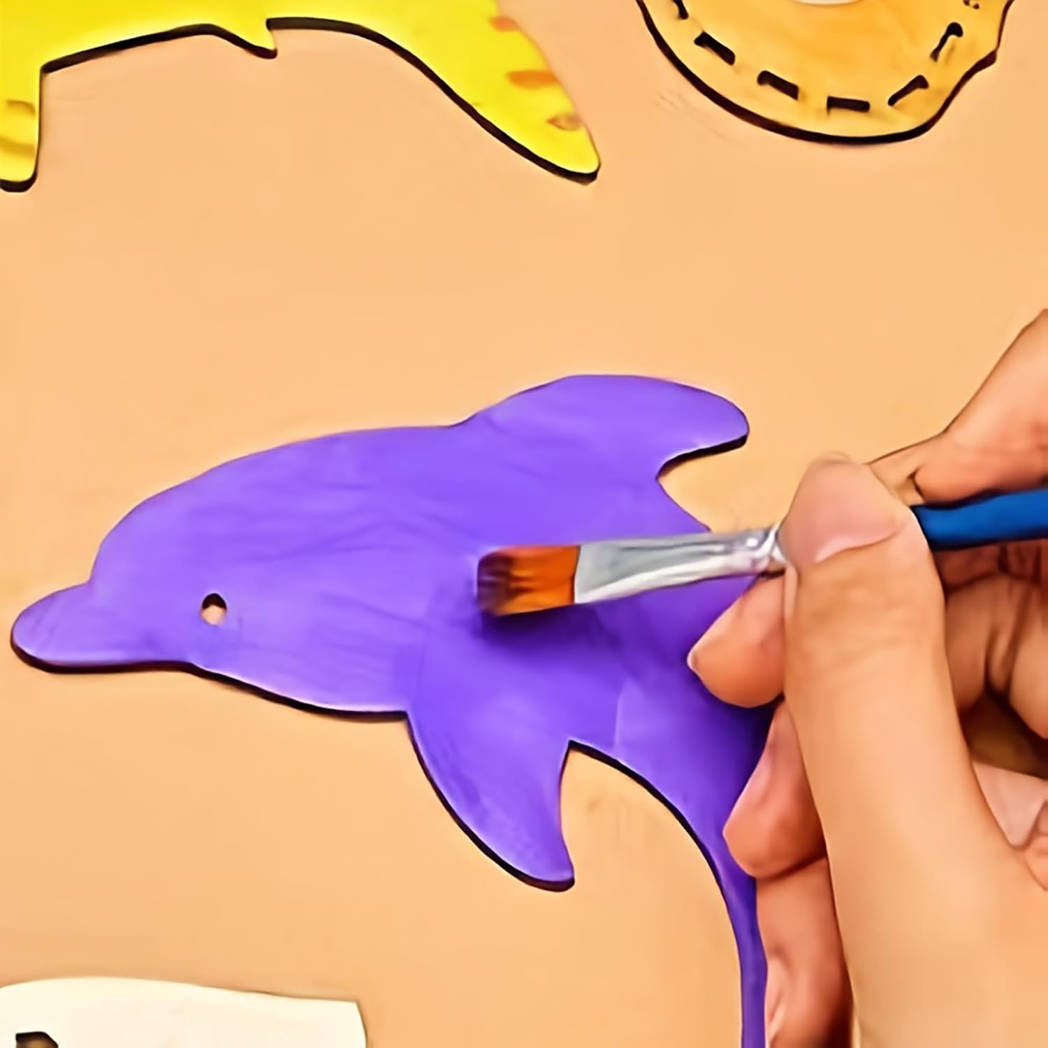 Hand painting a purple dolphin shape on a beige surface with a yellow dinosaur outline.