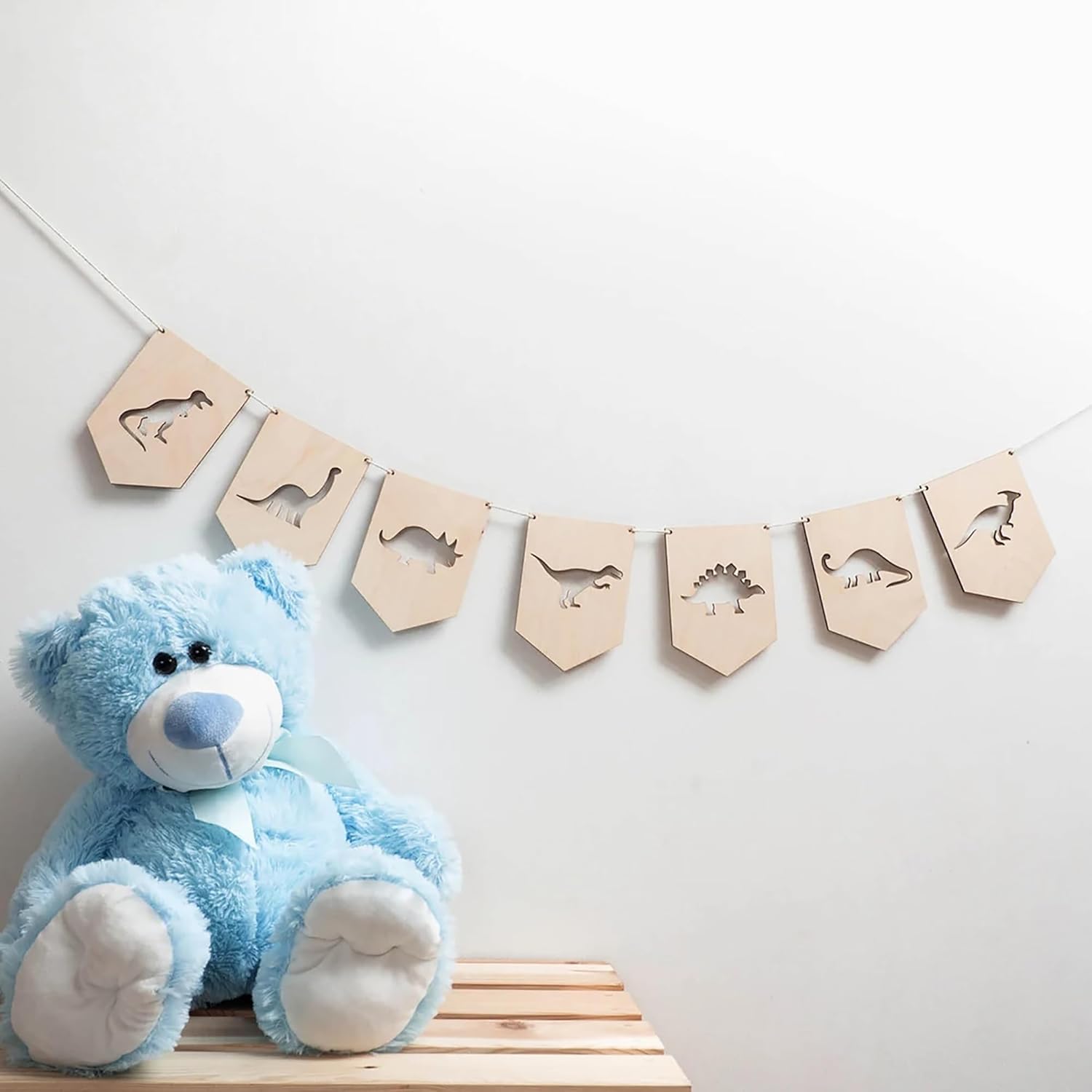 Blue teddy bear next to a wooden banner with dinosaur shapes on a light gray background