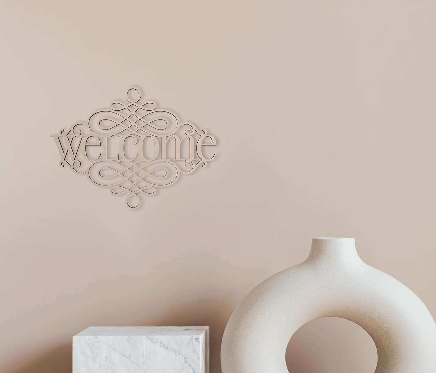 Decorative 'welcome' wall art on a beige wall with a white vase and box below.
