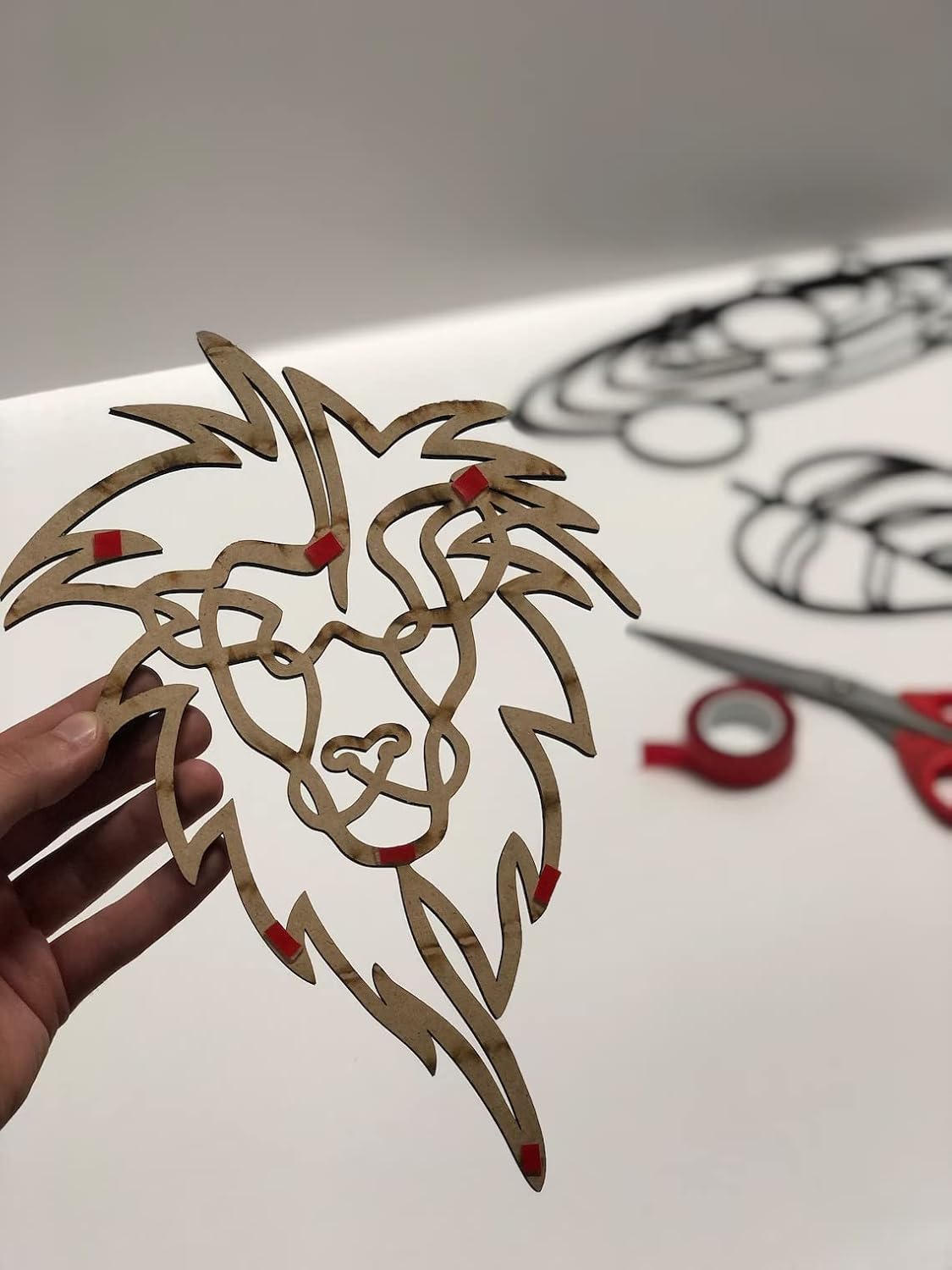 Hand holding a cut-out paper design of a lion's head with red accents on a white surface.