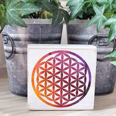 Square tile with a colorful flower of life design on a wooden surface with plants in the background