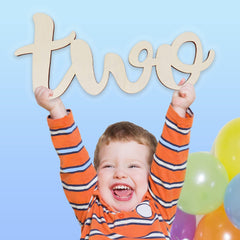 Child holding up a wooden 'two' sign with balloons in the background