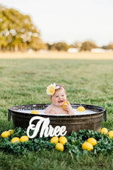 Baby in a tub with lemons and a 'Three' sign outdoors