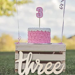 Pink cake on a wooden crate with 'three' spelled out, outdoors.