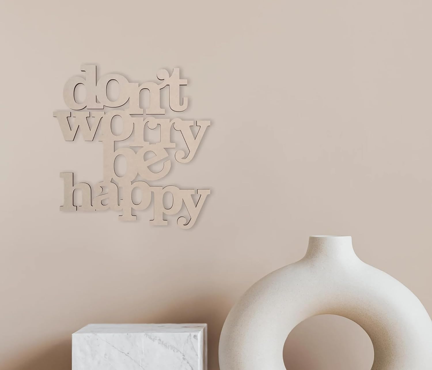 Decorative wall art with 'don't worry be happy' on a beige wall with a white vase and box.