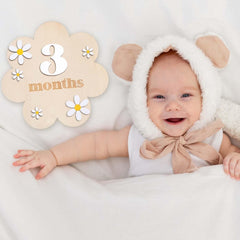 Baby in a white outfit with bear ears and a pink bow, lying on a white blanket next to a '3 months' milestone board.