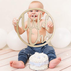 Baby sitting with a 'Wild One' sign and a small cake on a wooden floor.