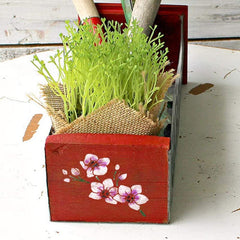 Red wooden planter with floral design containing green plants on a white surface.