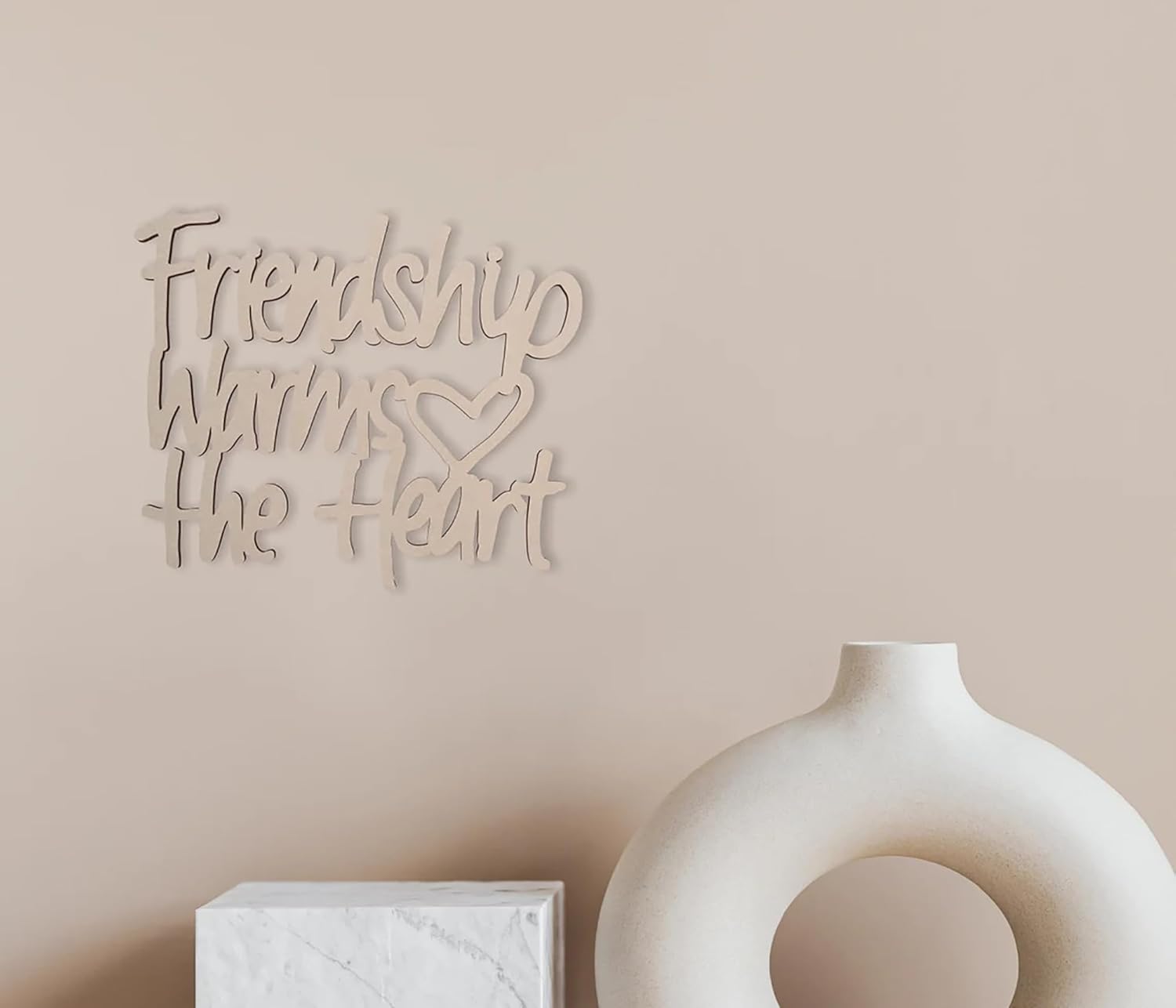 Decorative wall art with 'Friendship warms the heart' text on a beige wall with a vase and box below.