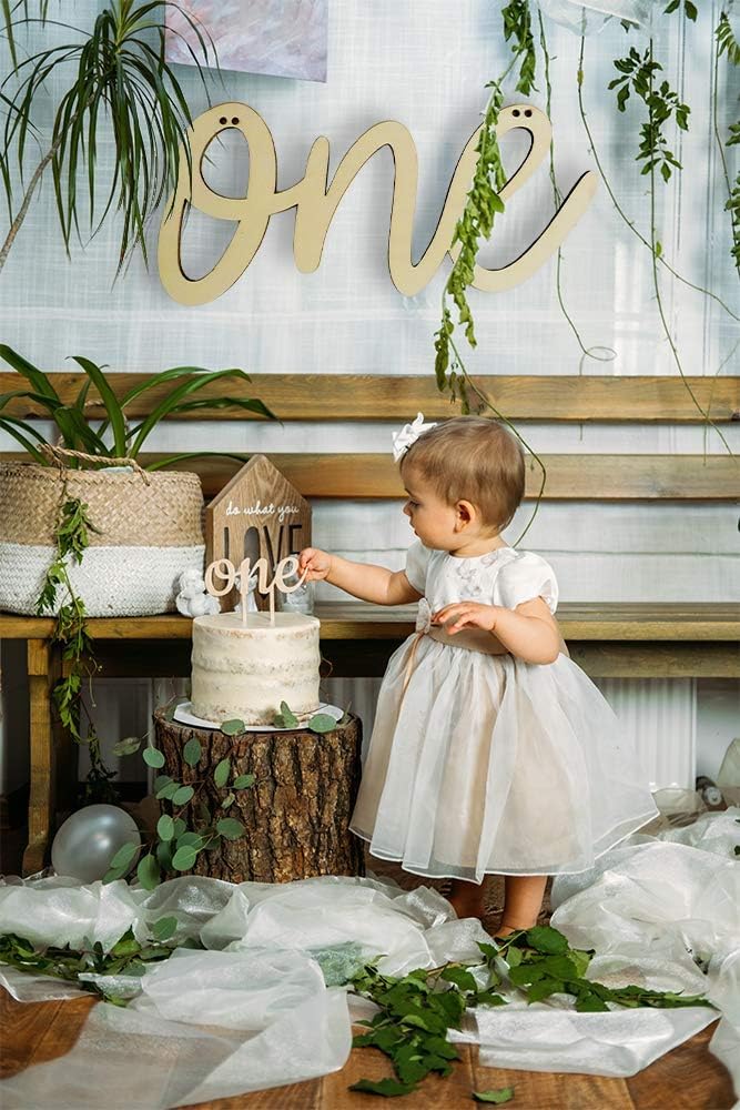 Child in a white dress standing next to a small cake with 'one' on it, surrounded by greenery and wooden decor.