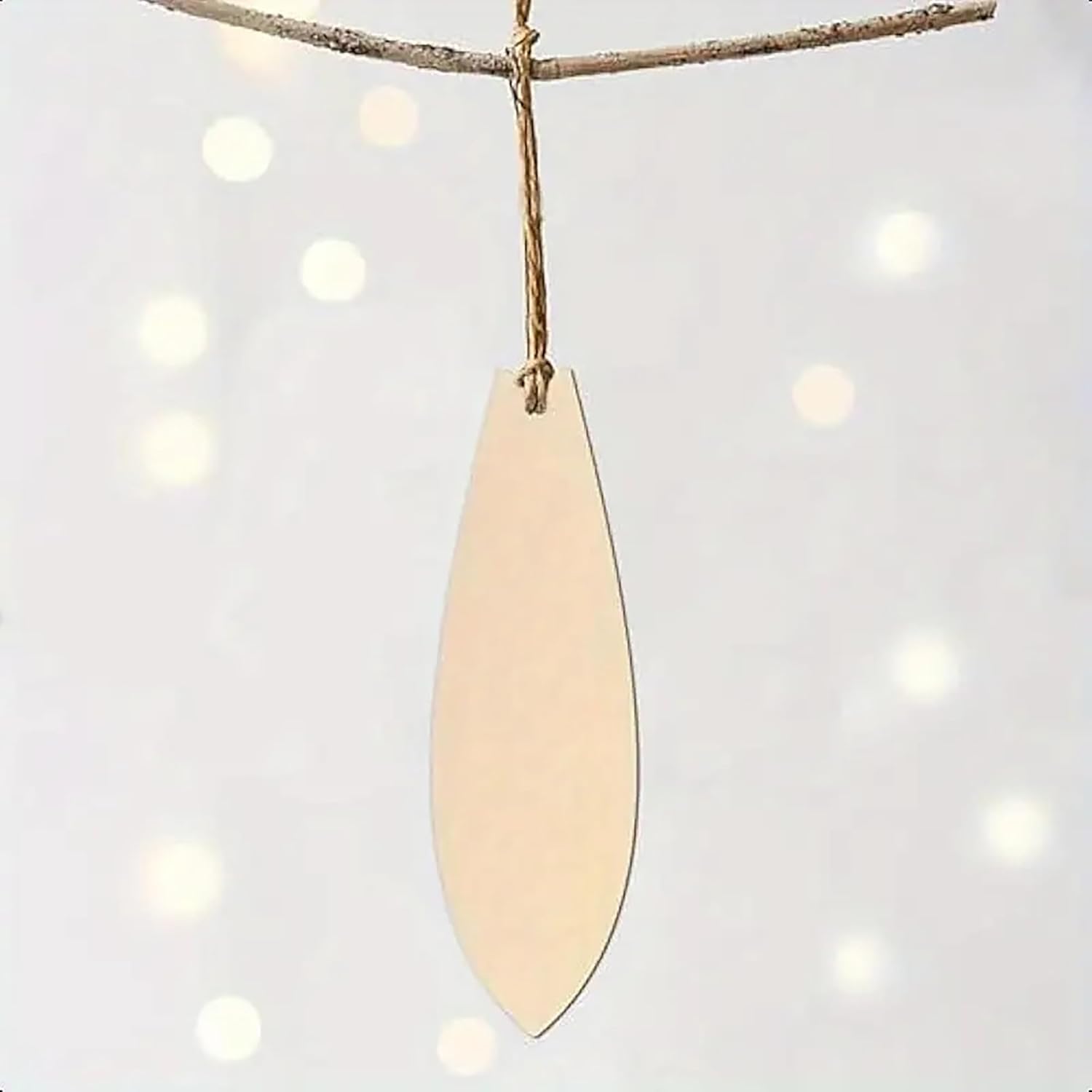 Beige teardrop-shaped ornament hanging from a branch against a white background with blurred lights.