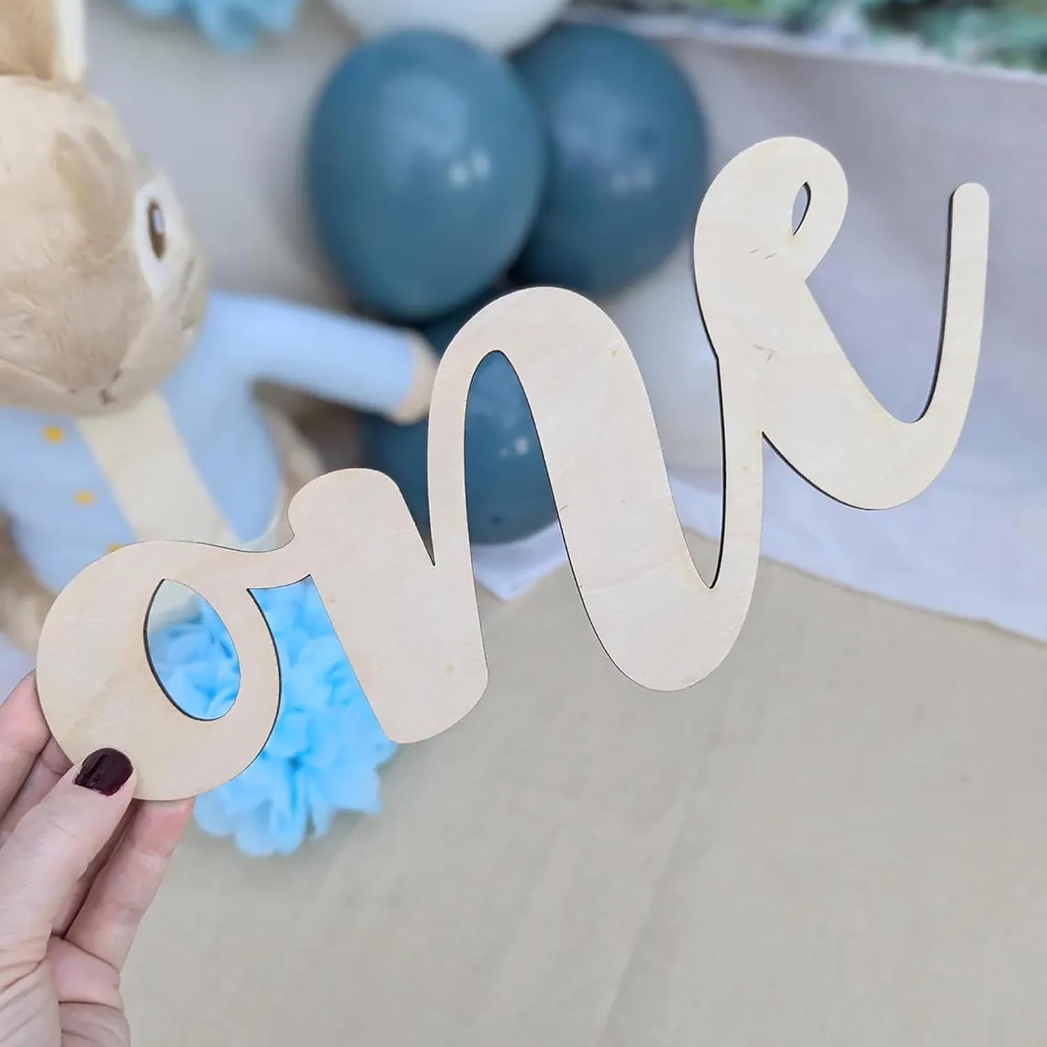 Wooden letter 'one' held by a hand with blue balloons and decorations in the background.