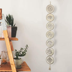 Decorative wall hanging with circular designs on a white wall next to a wooden shelf with plants.