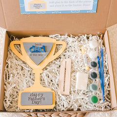 Craft kit with wooden plaque, paint bottles, and brushes in a box labeled 'Happy Father's Day'.