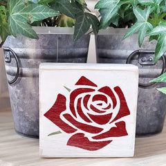 Wooden block with red rose design in front of metal buckets and green plants