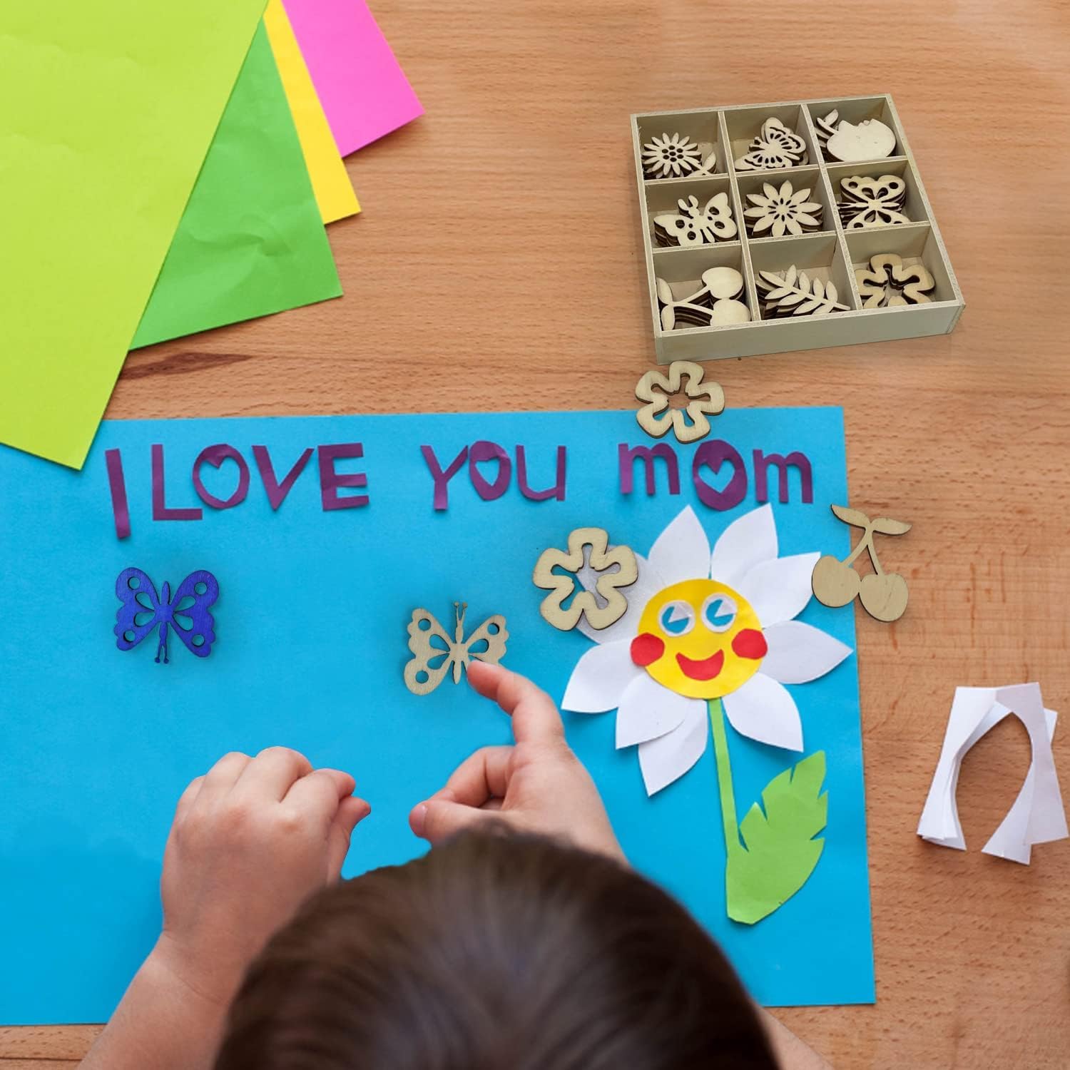 Child making a 'I Love You Mom' card with paper cutouts on a wooden table.