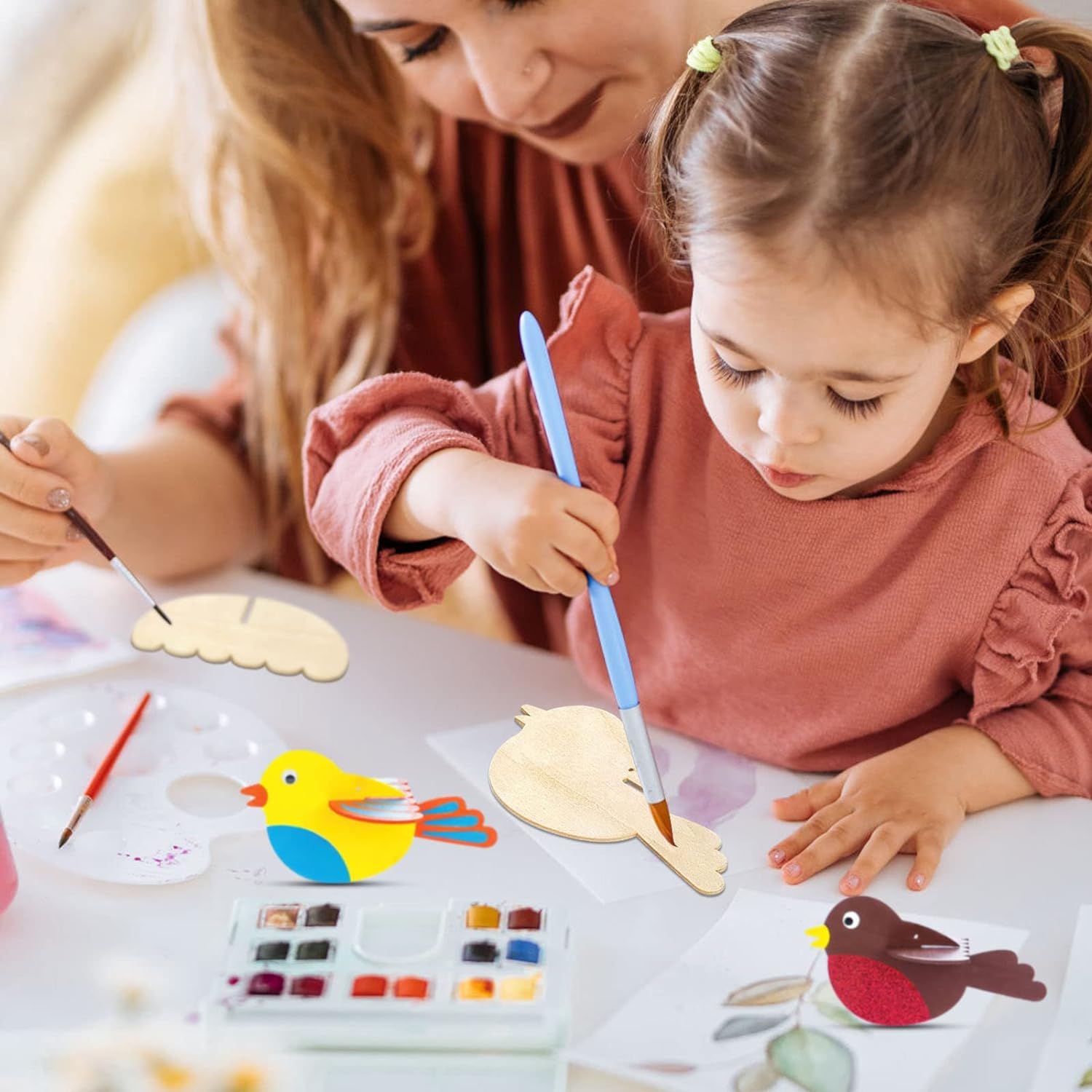 Child painting wooden craft items with a woman assisting, surrounded by art supplies.